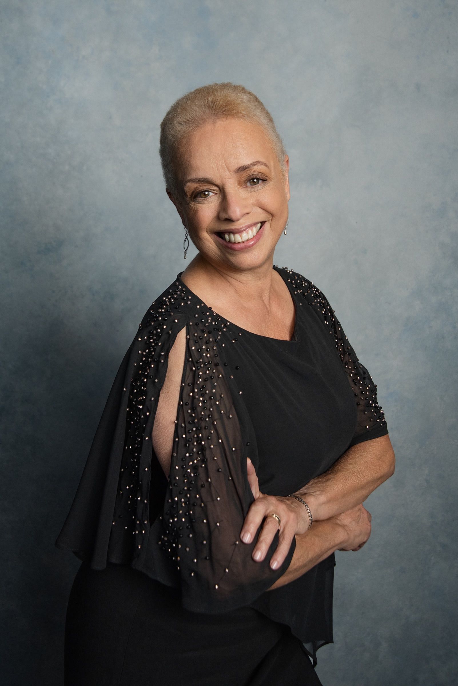 A woman in her sixties with cropped platinum hair and a wide smile, hand on hip in a black beaded top.