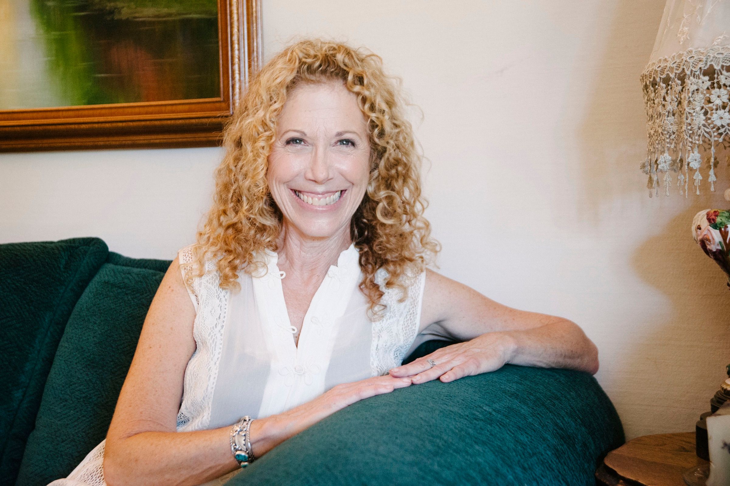 A woman in her fifties with curly blonde hair, smiling, leaning on a green sofa in a white lace blouse.