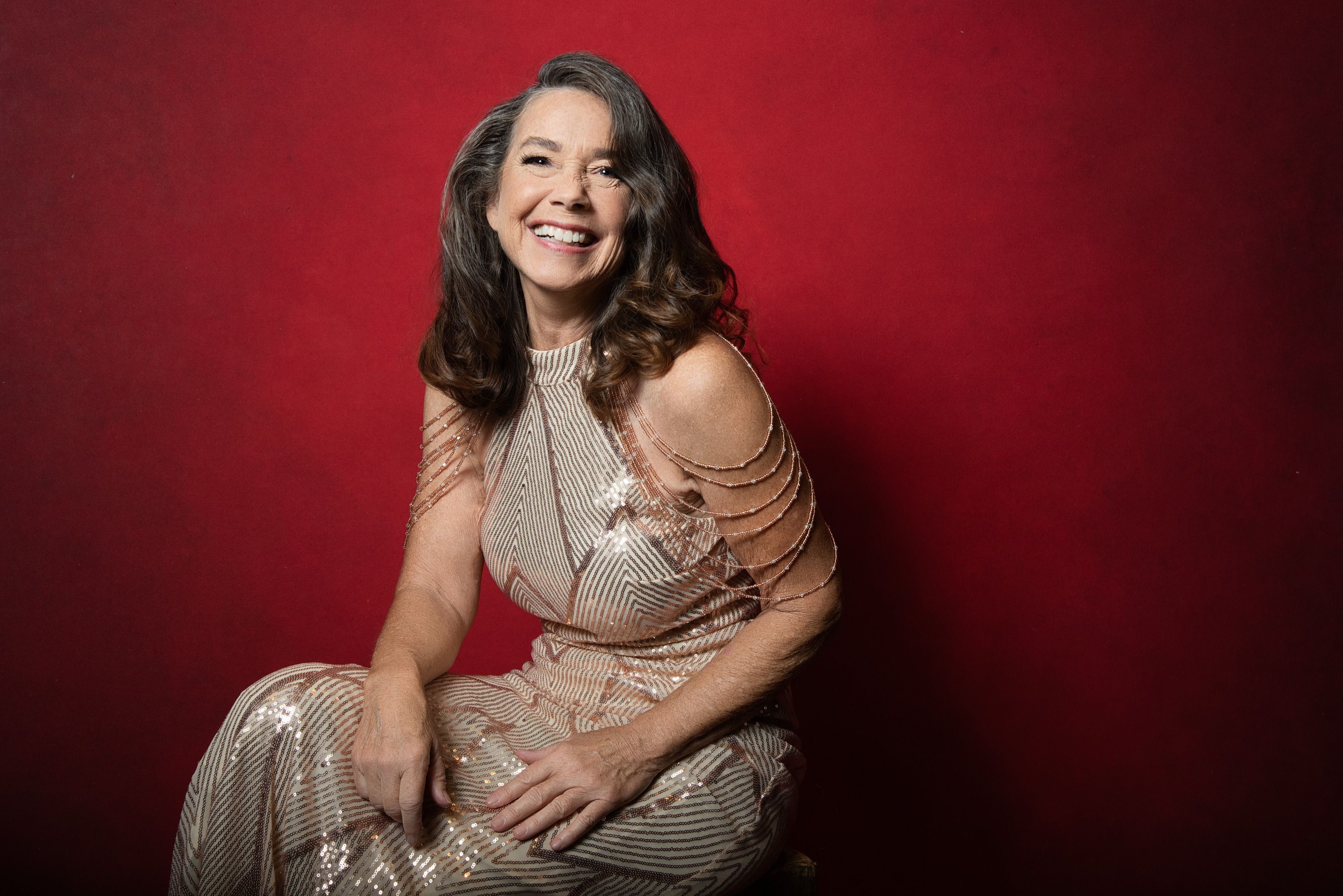 A woman in her fifties with dark shoulder-length hair, laughing, seated in a champagne beaded gown against a deep red backdrop.