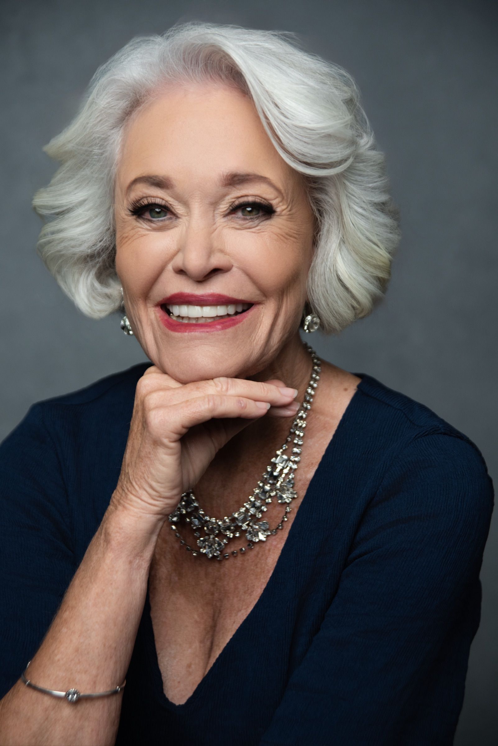 A woman in her seventies with full silver hair and a crystal necklace, chin resting on her hand, smiling broadly.