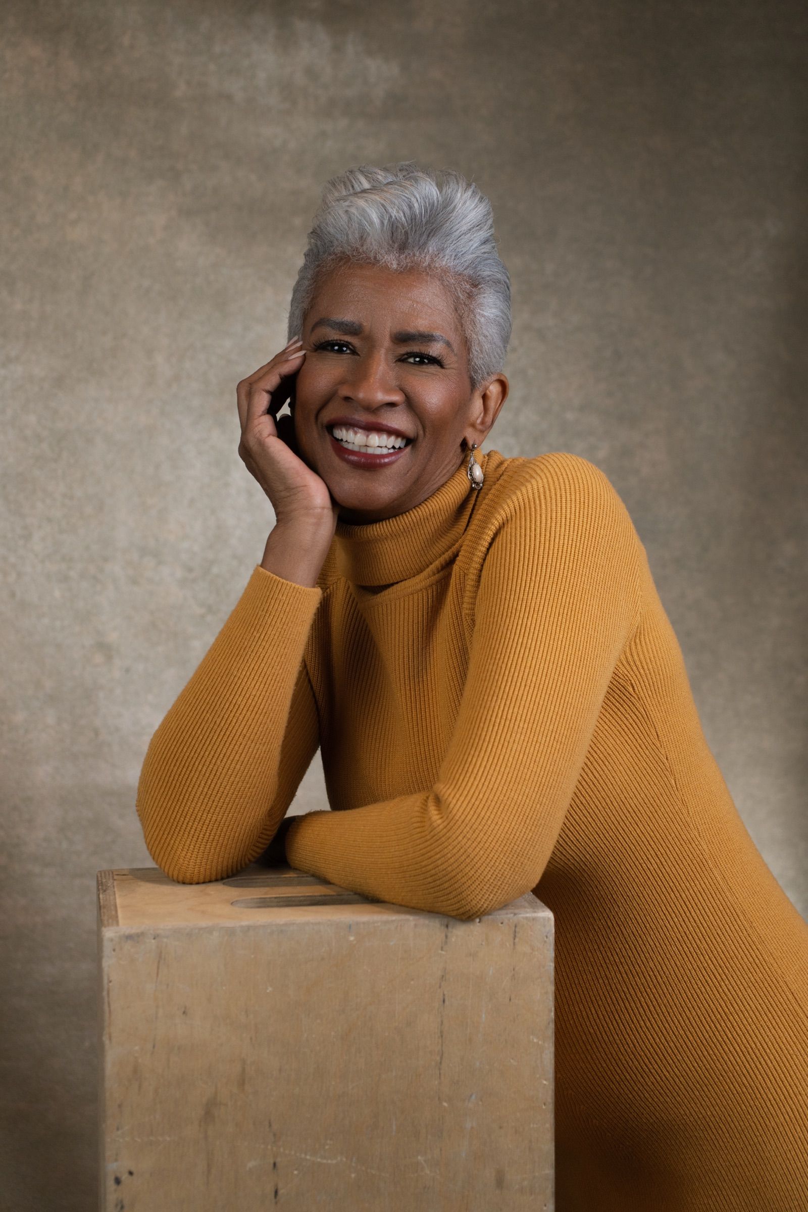 A woman in her sixties with a silver pixie cut, smiling with her hand at her cheek, leaning on a wooden box in a mustard turtleneck.